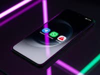 Un smartphone affichant trois icônes d'applications de messagerie : Telegram, WhatsApp et Signal sur une surface réfléchissante avec des lumières néon.