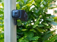 Une caméra de sécurité Blink Arc montée sur un poteau en bois, entourée de feuillage vert luxuriant.