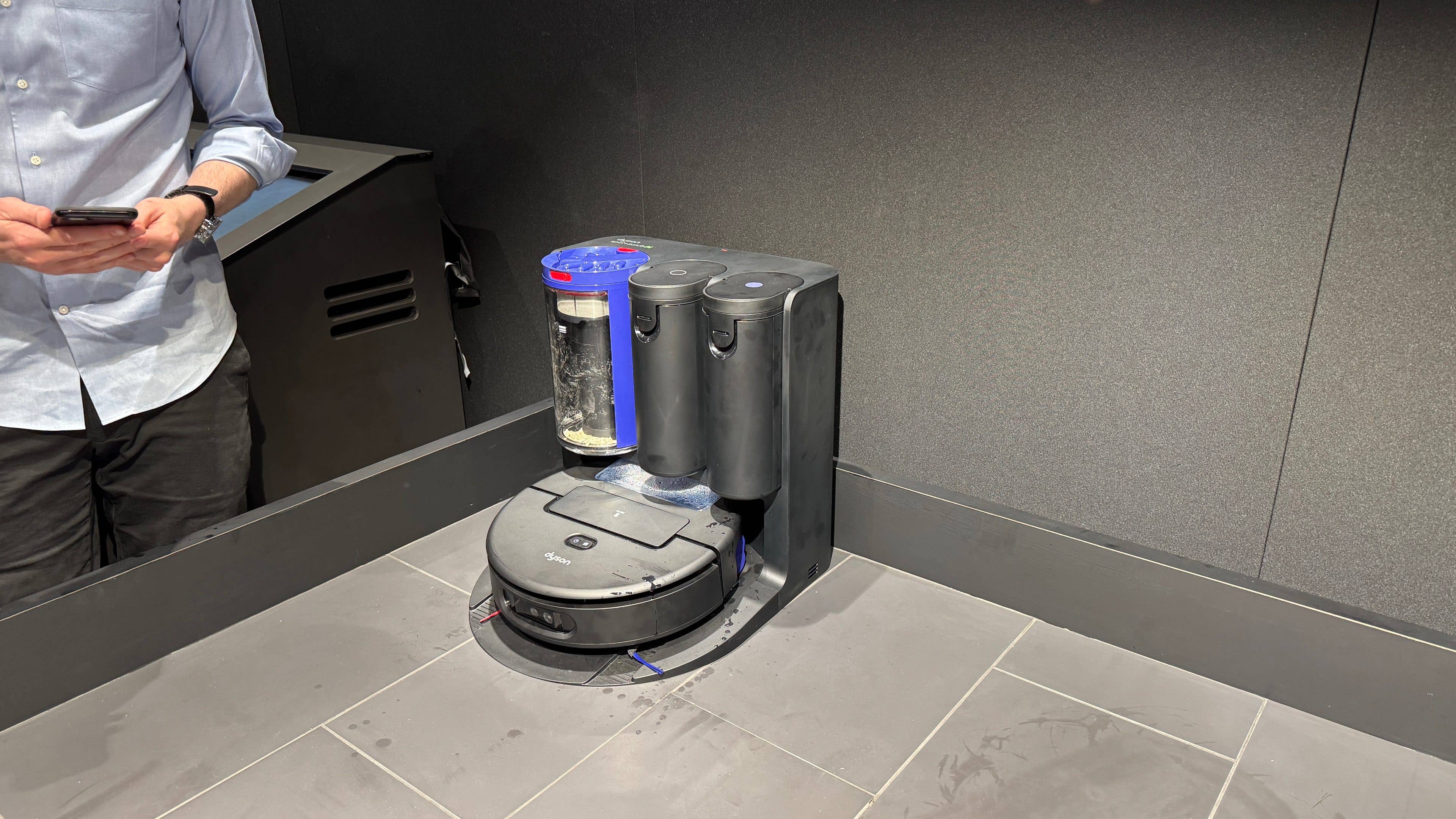 Un nettoyeur robot Dyson Spot+Scrub Ai sur un sol carrelé, avec une personne tenant un téléphone à proximité.