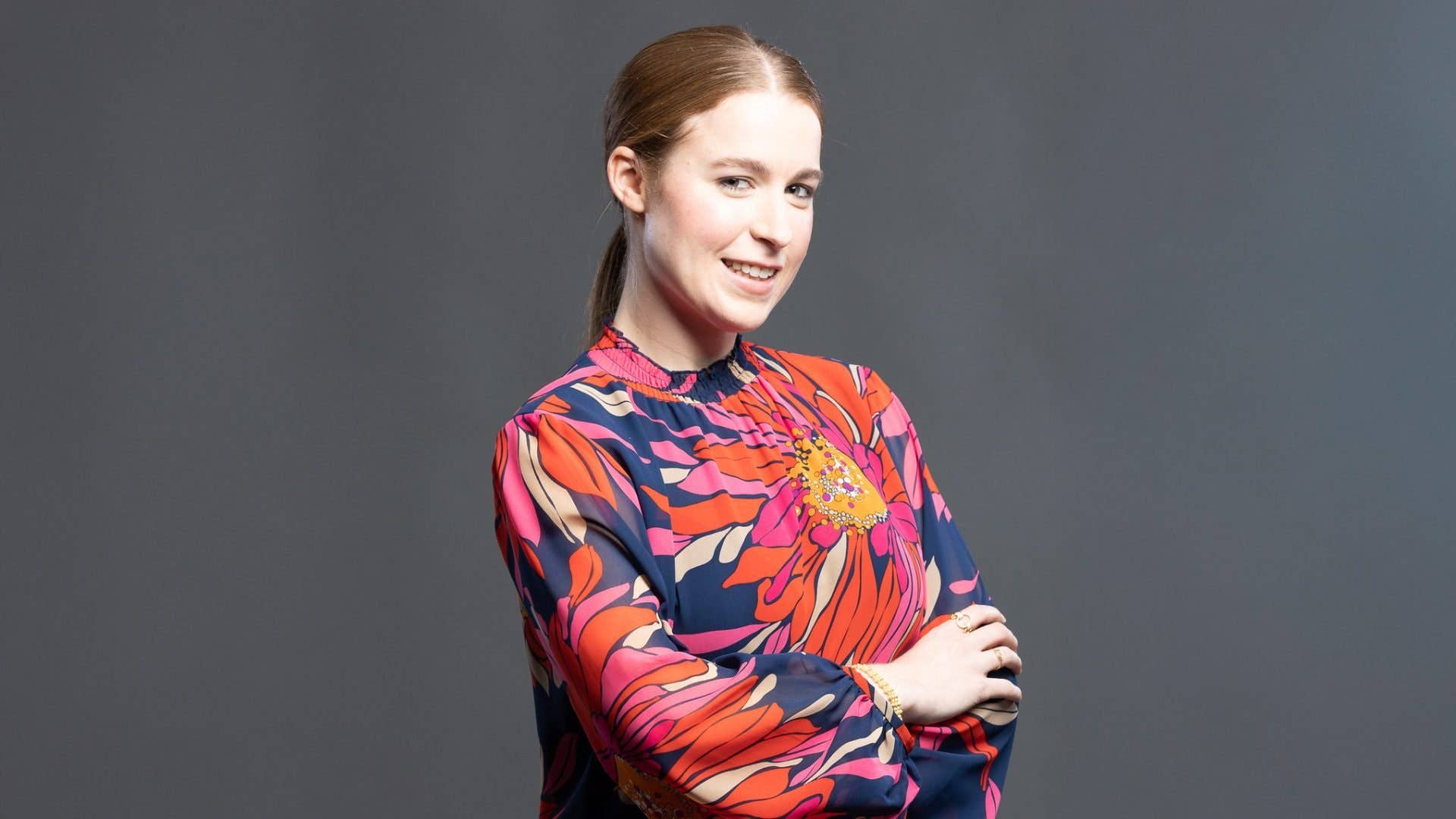 Une femme portant un chemisier floral coloré sourit et pose avec assurance, bras croisés.