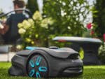 Un robot de tonte Gardena Smart Sileno Sense sur de l'herbe verte avec une personne se relaxant en arrière-plan.