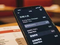 Un smartphone affichant un menu avec des plats comme le Kung Pao Chicken et le Mapo Tofu, avec les prix dans un restaurant.