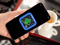 Une main tenant un smartphone affichant le logo Android 16 sur une table avec une tasse de café.