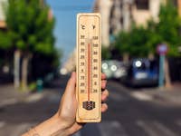 Une main tenant un thermomètre en bois affichant des températures élevées en Celsius et Fahrenheit dans une rue ensoleillée.