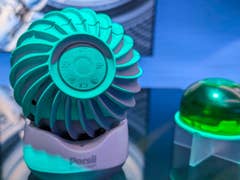 Un appareil vert en forme de ventilateur étiqueté 'Persil smartwash' à côté d'une capsule gélifiée verte sur une surface réfléchissante.