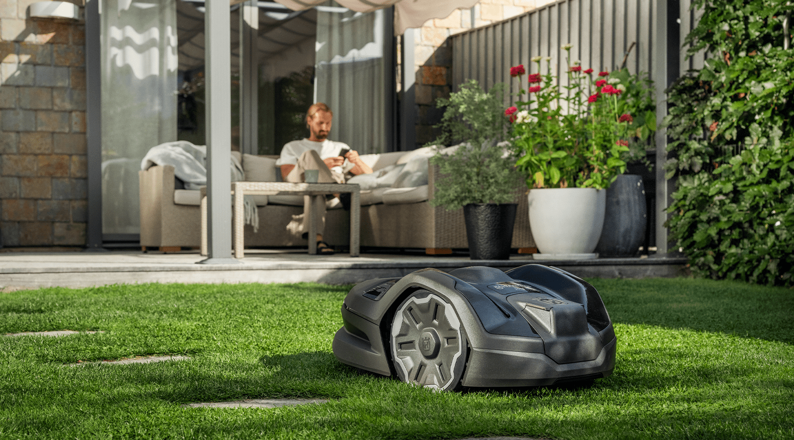 Une tondeuse à gazon robot Husqvarna 305XE Nera sur une pelouse verte avec un homme se relaxant sur un patio en arrière-plan.