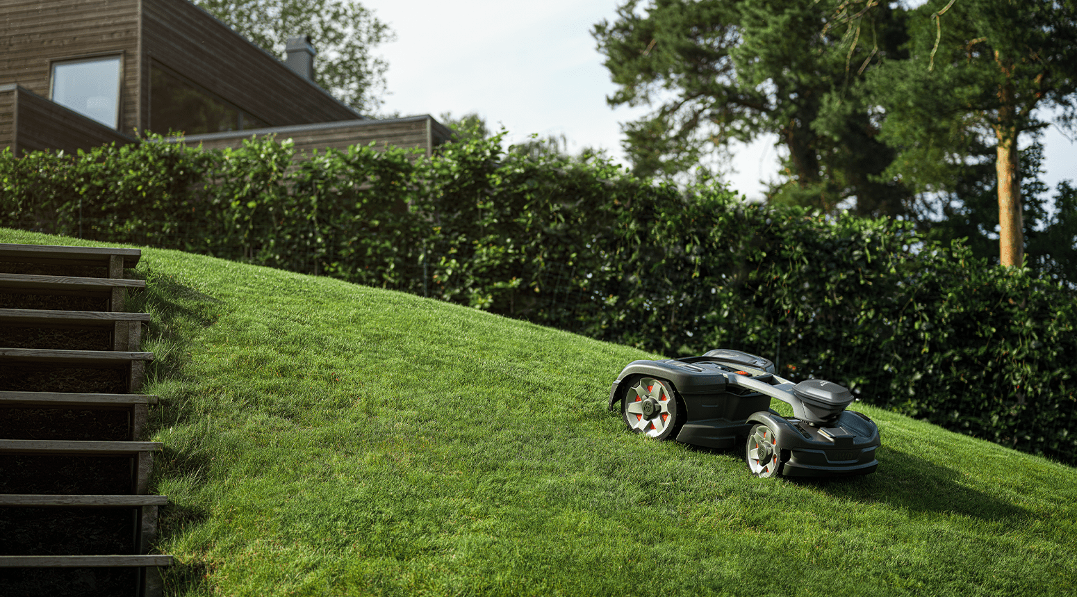 Une tondeuse à gazon robotisée Husqvarna sur une pelouse en pente à côté d'une maison en bois moderne.