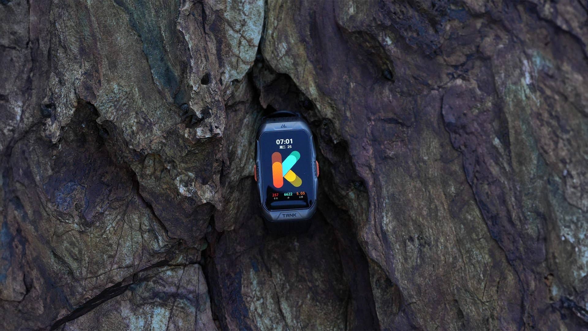 Une montre intelligente Kospet Tank X2 affichée sur une surface rocheuse, montrant l'heure 07:01 et un logo coloré.