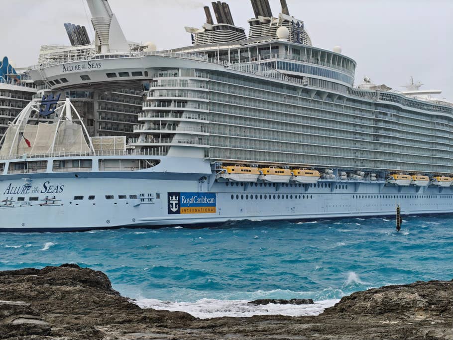 Un grand paquebot de croisière, Allure of the Seas, amarré près de rivages rocheux avec des eaux bleues au premier plan.
