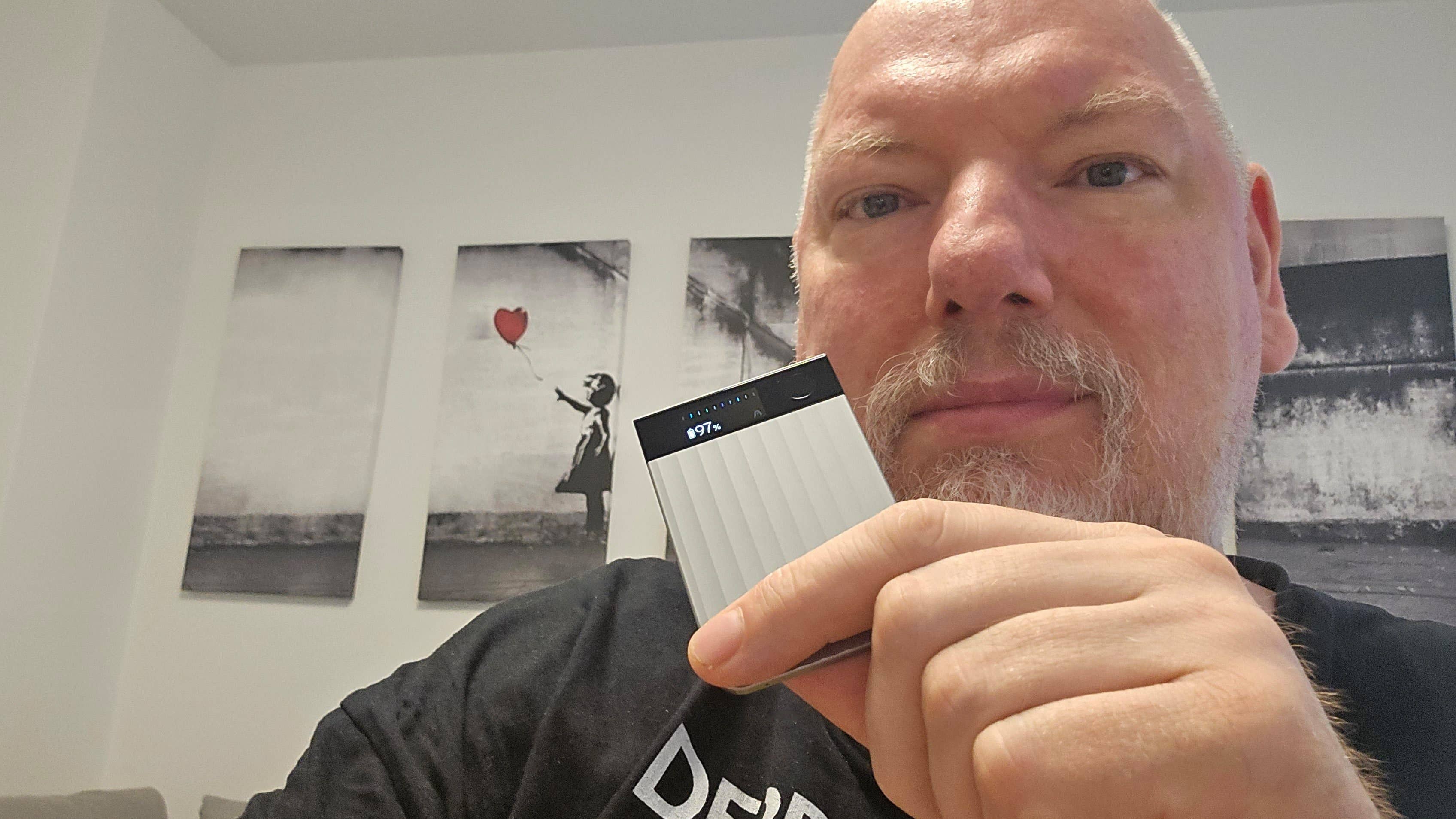 Plaud_Note_Pro_Hero.jpg Une personne tenant un petit appareil étiqueté '97%' devant une œuvre d'art représentant une fille avec un ballon.