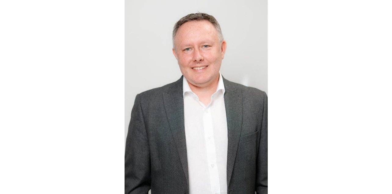 Un homme souriant en costume gris et chemise blanche, se tenant devant un fond clair.