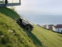 Roborock RockMow Z1 tondant de l'herbe sur une pelouse inclinée près d'une maison moderne surplombant la mer.