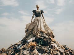 Un mannequin portant un haut noir et une grande jupe beige se tient au sommet d'un tas de vêtements usagés sous un ciel nuageux.