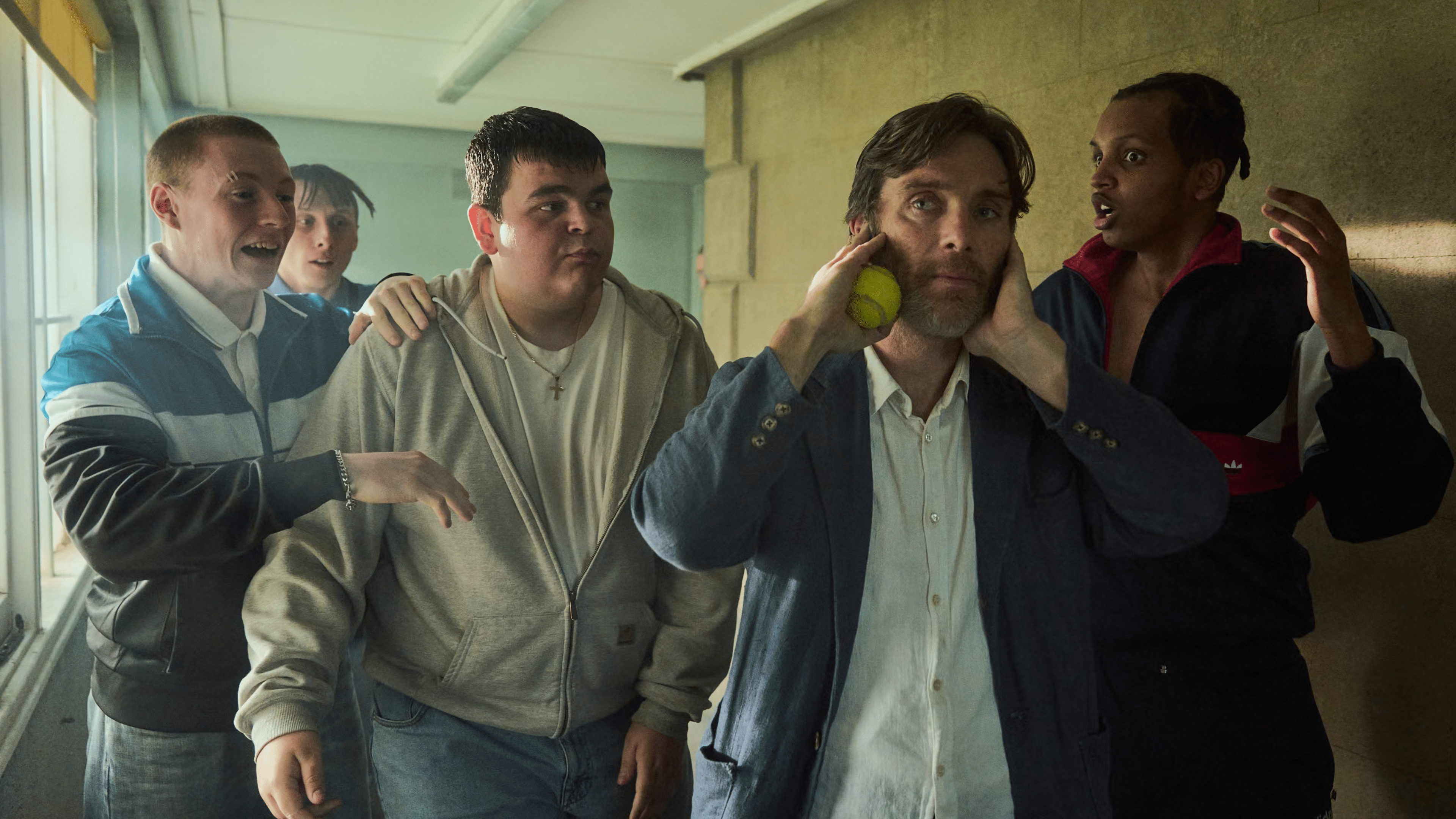 Un groupe de quatre jeunes hommes entoure un homme inquiet tenant sa tête, tandis qu'un lance une balle de tennis.