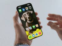 Un iPhone vu de face, tenu dans une main gauche, avec son écran allumé affichant l'écran d'accueil d'iOS 18.