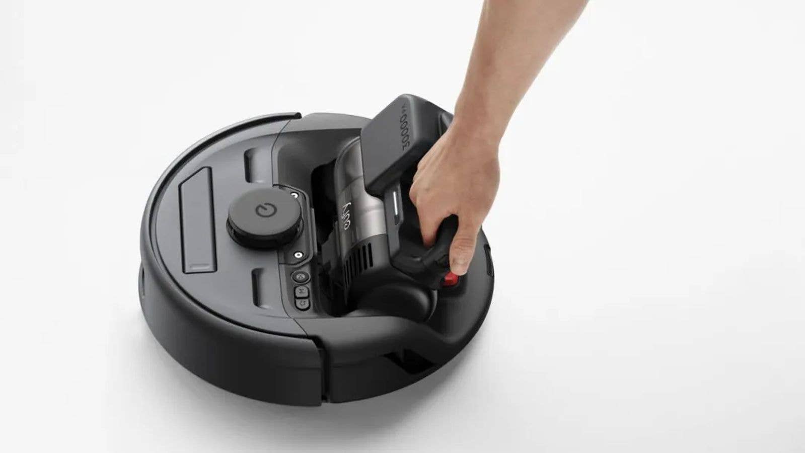 Une personne tient un aspirateur à main au-dessus d'un aspirateur robot sur une surface blanche.