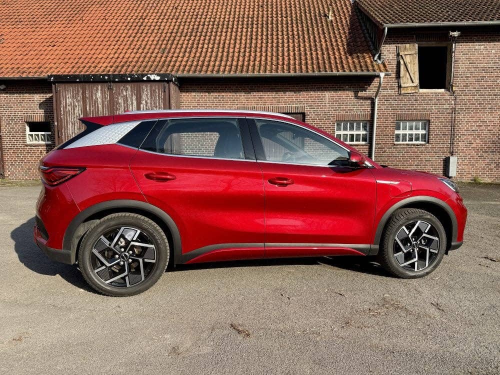 Vue latérale d'une BYD Atto 3 rouge, garée sur une surface de gravier près d'un bâtiment en briques.