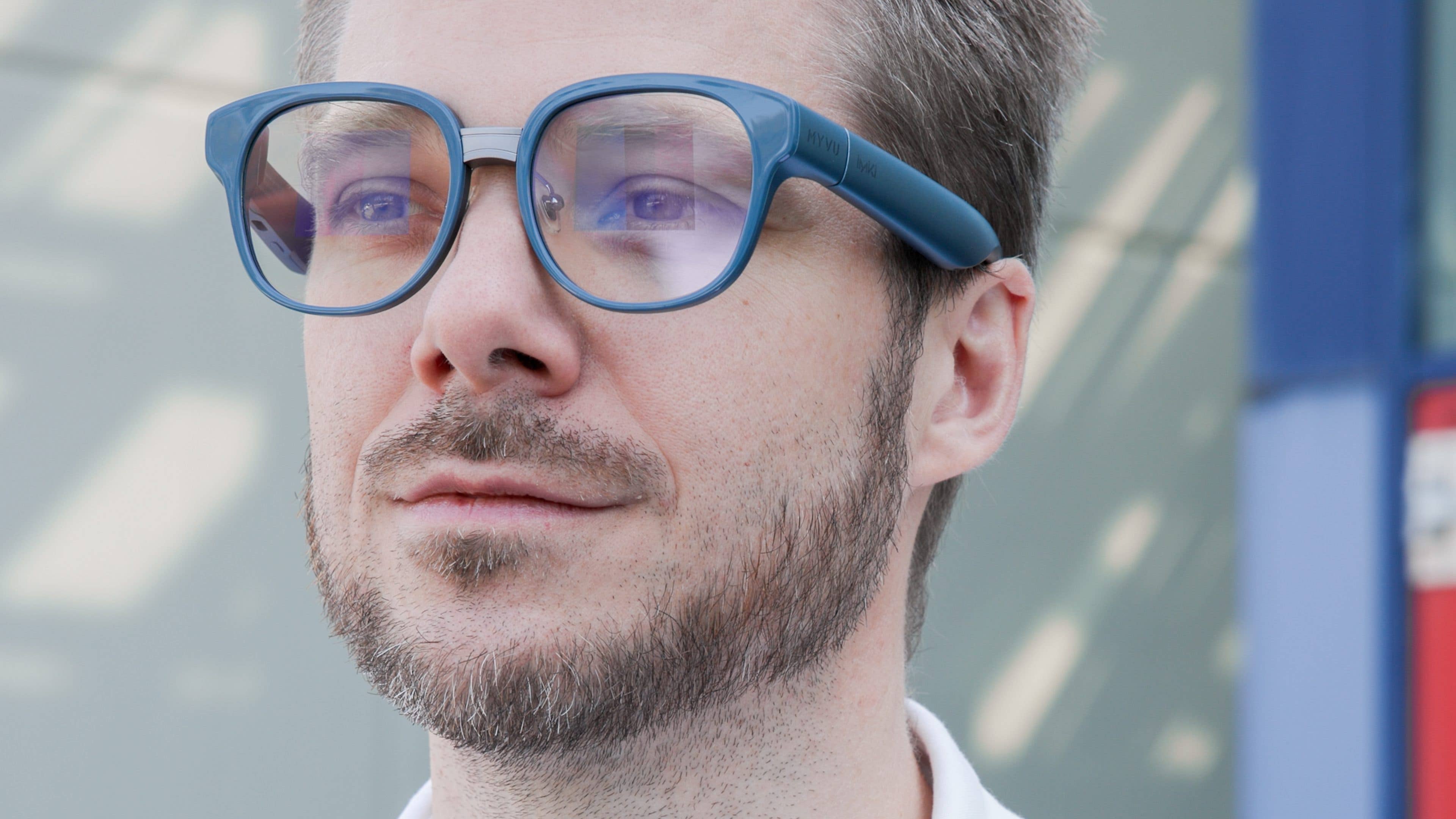Un homme avec une barbe claire portant des lunettes bleues, regardant pensivement au loin.
