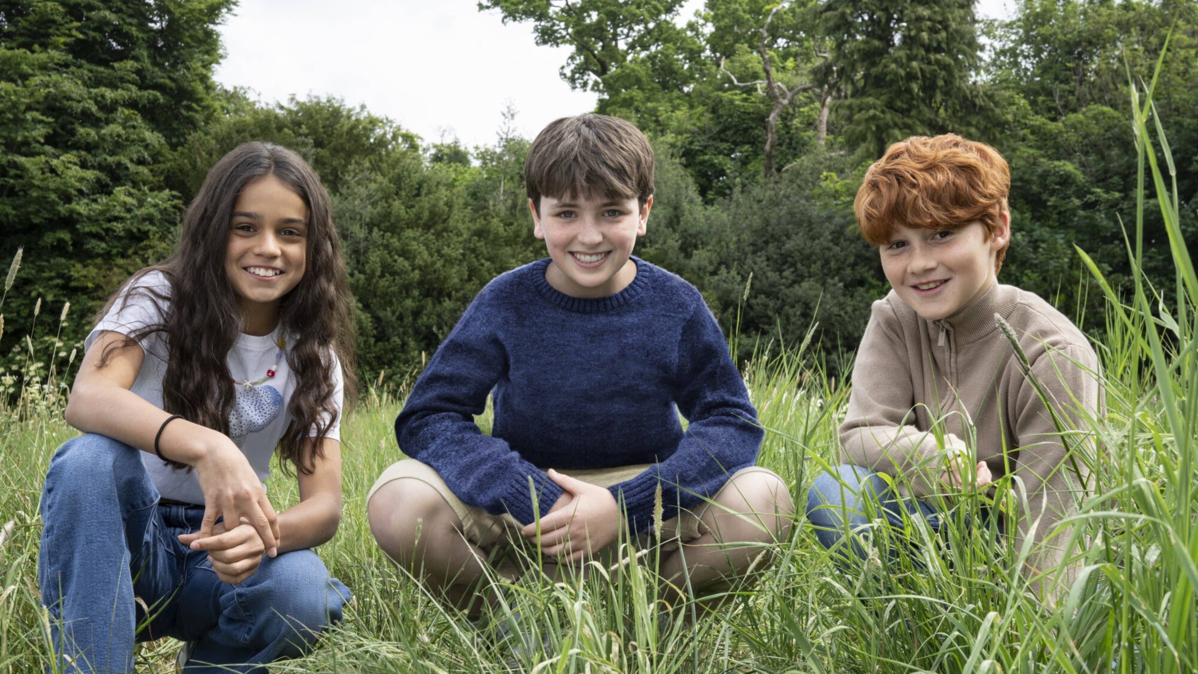 Trois enfants souriant assis dans l'herbe, avec des arbres en arrière-plan.