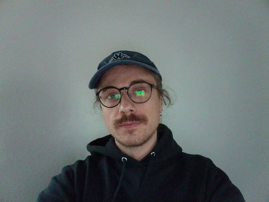 Une personne avec des lunettes et une moustache, portant une casquette et un sweat à capuche devant un mur gris.