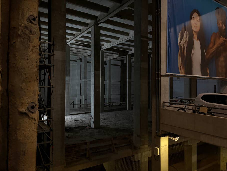 Un chantier de construction vide éclairé par une seule source de lumière, avec des piliers en béton et une grande bannière publicitaire.