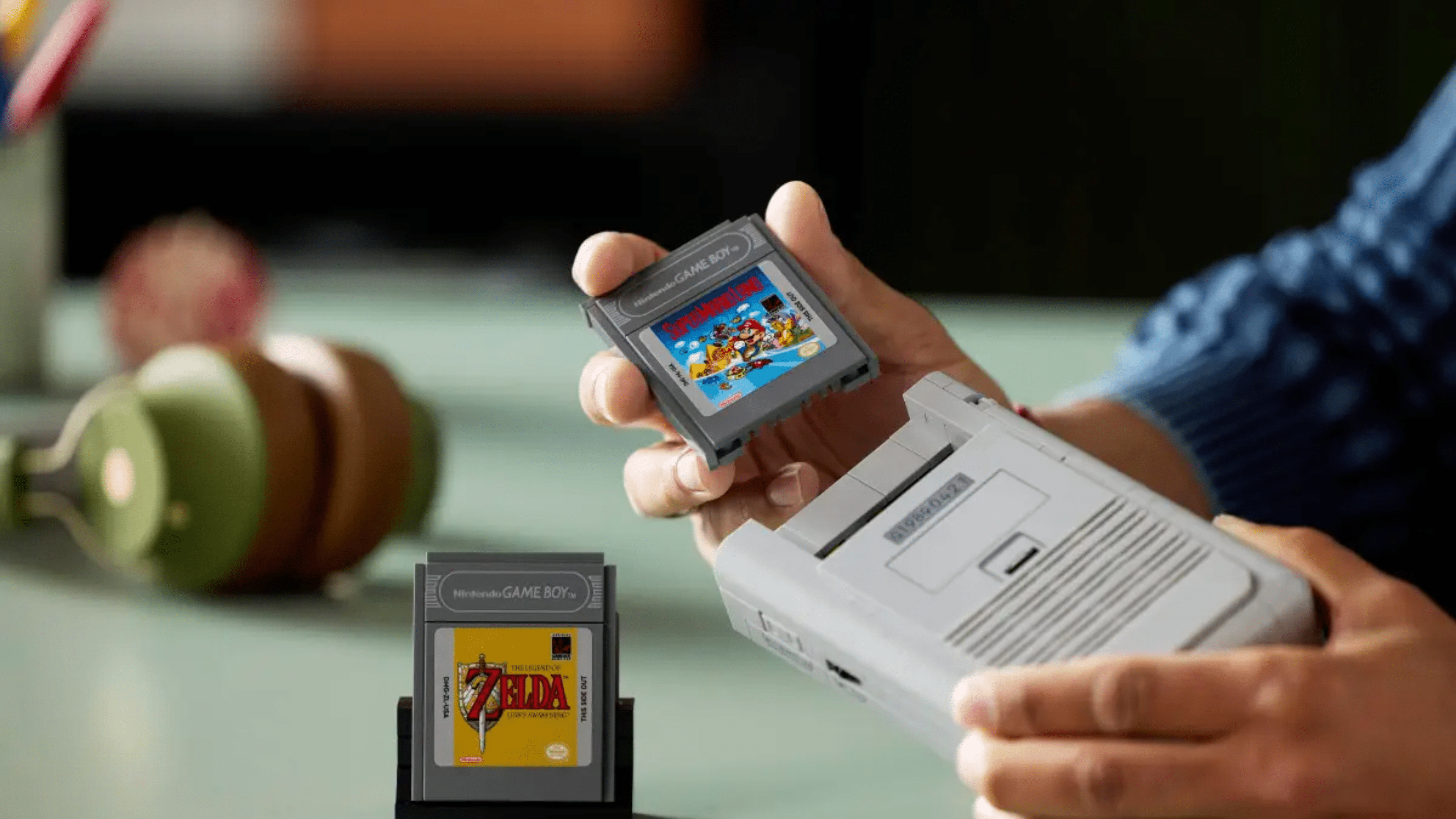 Une personne tient une cartouche de Game Boy étiquetée 'Super Mario' au-dessus d'une console Game Boy avec la cartouche 'Zelda'.