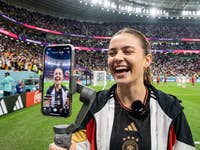Une créatrice de contenu se tient au bord du terrain dans un stade et parle à la caméra de son téléphone portable.
