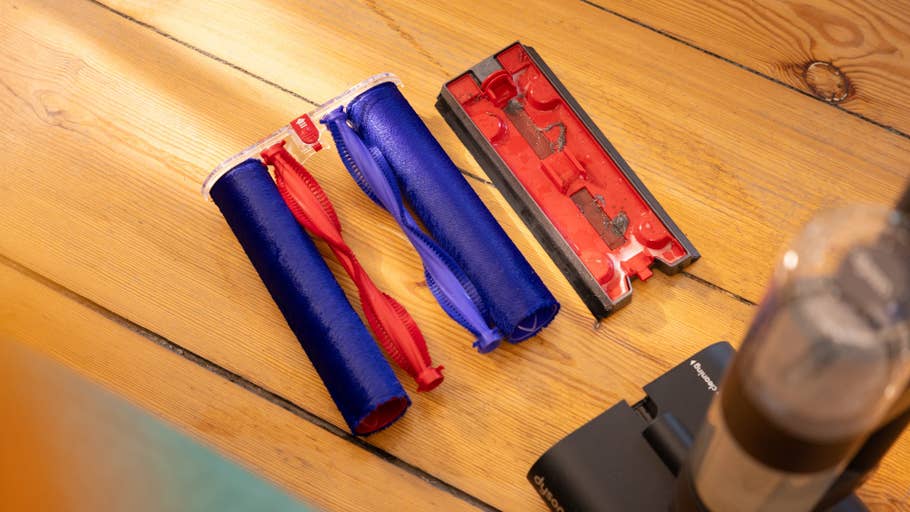 Pièces du0027aspirateur Dyson : deux rouleaux de brosse (bleu et rouge) et un plateau à débris rouge sur un sol en bois.