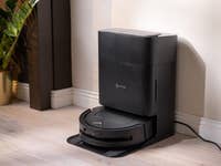 Un aspirateur robot Ecovacs T50 Omni noir à côté d'une plante contre un mur.