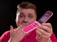 Une personne en chemise rouge tient un téléphone Barbie rose avec un écran coloré, se couvrant la bouche en jouant.
