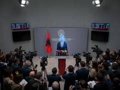 Une conférence de presse en Albanie avec un intervenant au pupitre, entouré de caméras et d'un drapeau albanais.