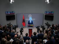 Une conférence de presse en Albanie avec un intervenant au pupitre, entouré de caméras et d'un drapeau albanais.