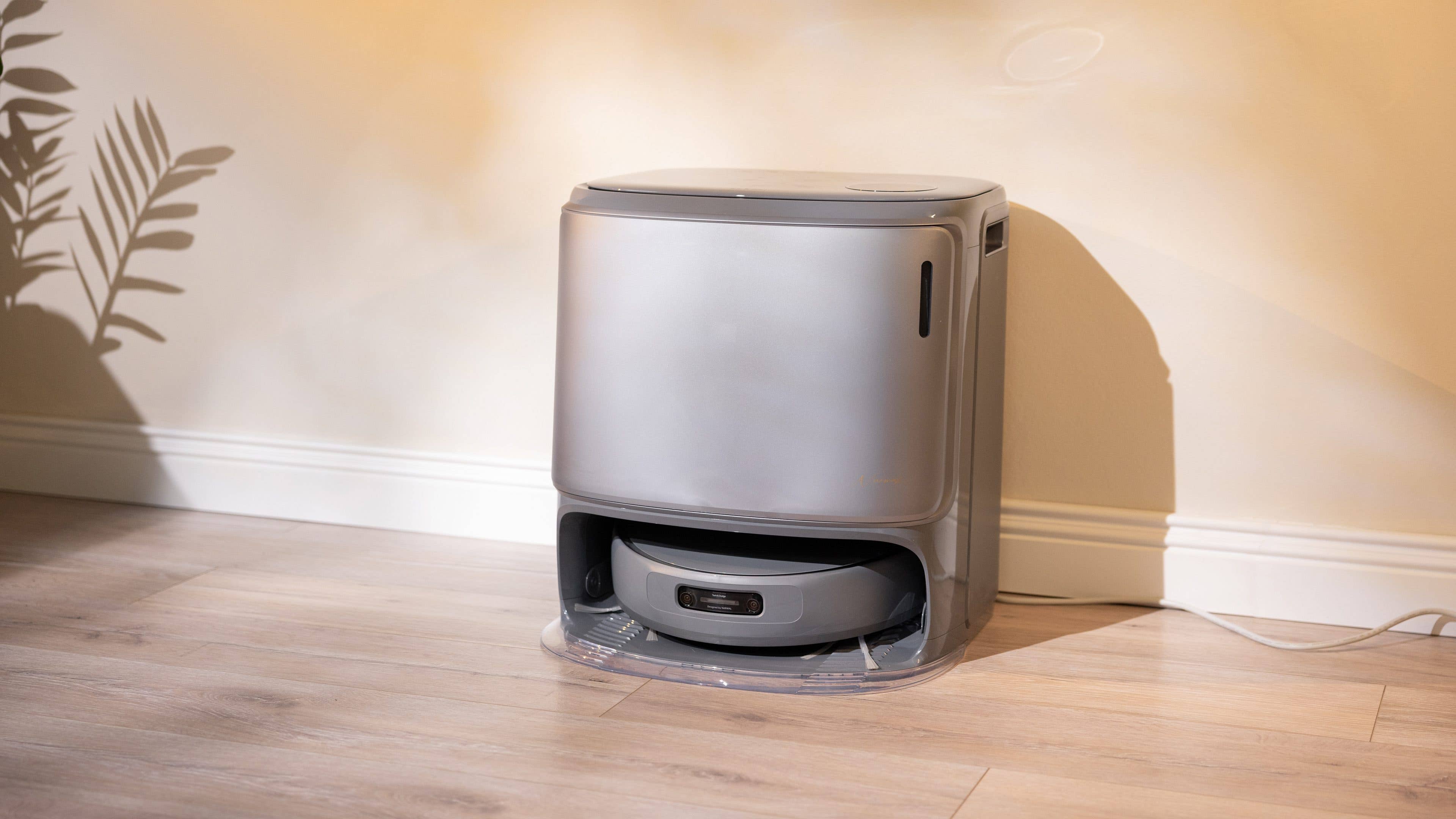 nextpit_Narwal_Z10_Ultra_Station.jpg Un aspirateur robot gris élégant se trouve contre un mur dans une pièce bien éclairée.