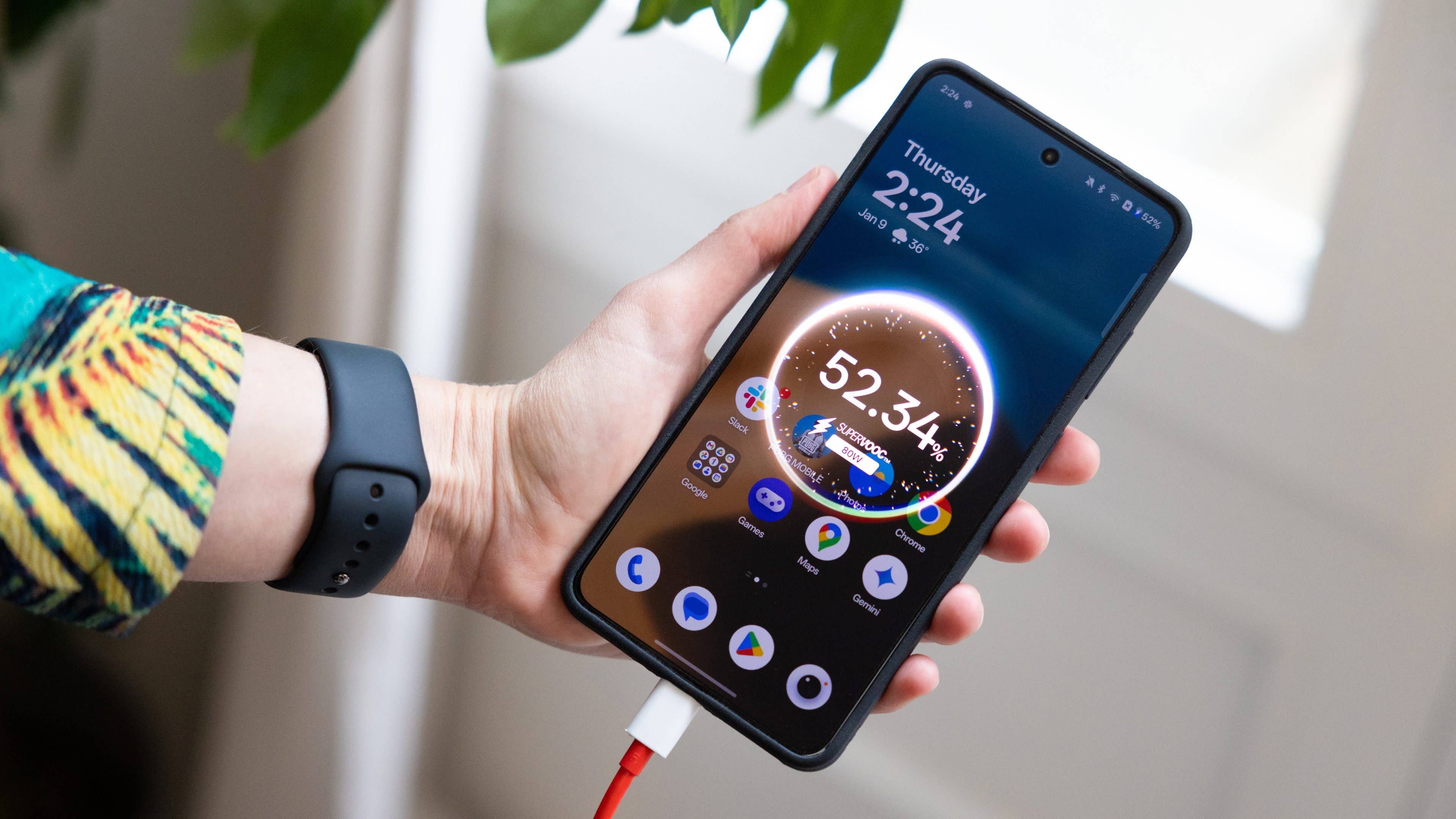 Une personne tient un smartphone OnePlus 13R affichant l'heure et l'état de la batterie, branché à un chargeur.