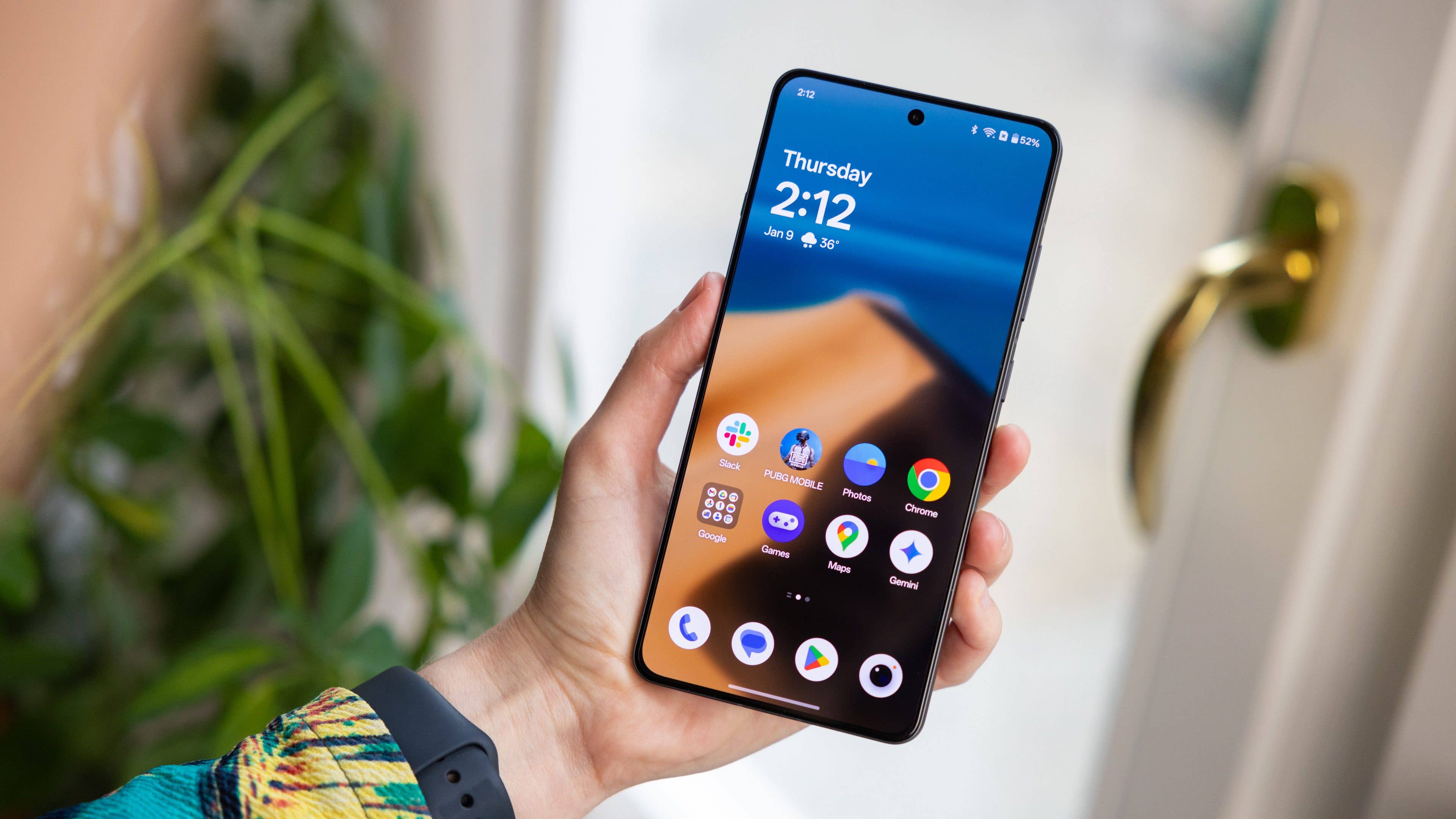 Une personne tenant un smartphone OnePlus 13R affichant l'heure 2:12 et diverses icônes d'applications.