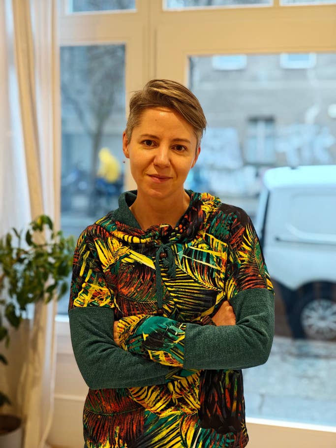 Une personne aux cheveux courts portant un sweat à capuche coloré se tient les bras croisés dans une pièce lumineuse.