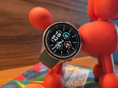 La OnePlus Watch 2R vue de de face avec son écran allumé et posée diagonalement contre une petite statuette rouge.