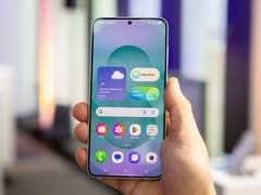 Une personne tenant un smartphone Samsung Galaxy S25 affichant un widget météo et des icônes d'applications.