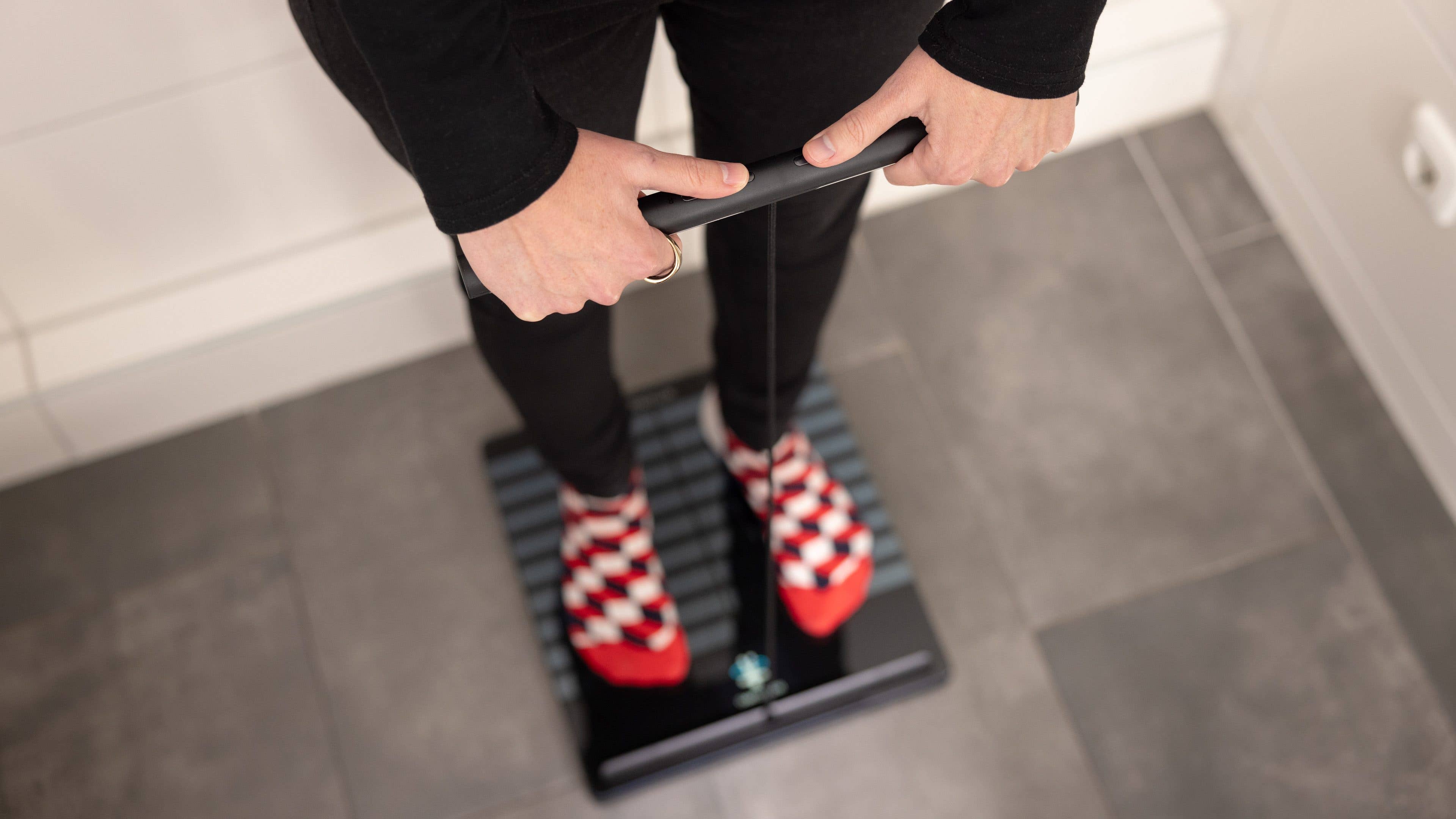 Personne debout sur une Withings Body Scan Smart Scale, tenant la poignée, portant des chaussettes à carreaux rouges et noires.