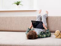 Enfant allongé sur un canapé, tenant une tablette, les pieds en l'air, à côté d'un chien en peluche.