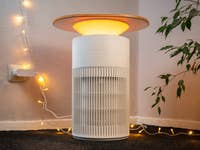 Un purificateur d'air SwitchBot avec un dessus en bois et une lumière douce, placé à côté d'une plante et de guirlandes lumineuses.