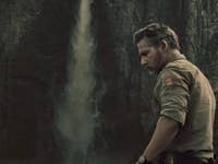 Un homme se tenant près d'une cascade, portant une chemise beige, avec une falaise rocheuse en arrière-plan.