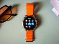 Une montre Xiaomi S4 avec un bracelet orange, affichant diverses icônes d'applications, à côté de lunettes de soleil et de magazines.